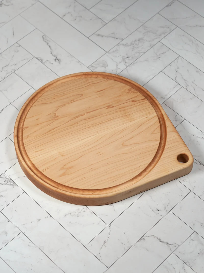 Hard maple round pizza cutting board with juice groove and grip hole, shown at an angle on white marble tile.