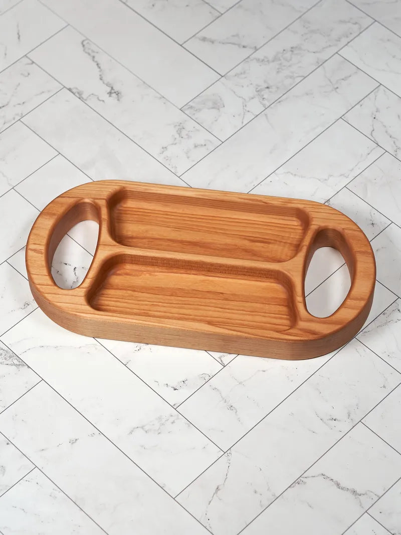 Hard maple divided serving tray with two chambers and oval handholds, shown at an angle on white marble tile.