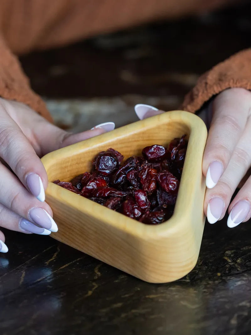 Hands holding a small maple wood snack bowl filled with dried cranberries on a dark surface
