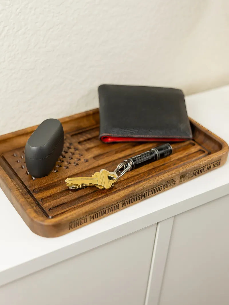 Walnut American flag tray on white counter holds keys with flashlight and black wallet. Hand holds black case above it.