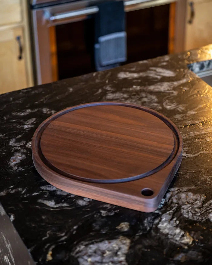 Empty walnut round pizza cutting board with juice groove on a black granite kitchen counter.