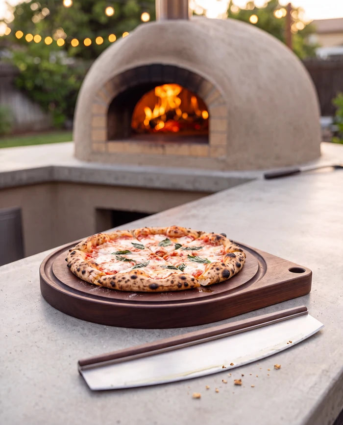 Fresh margherita pizza on a walnut round pizza board with a rocker cutter, in front of an outdoor wood-fired oven.