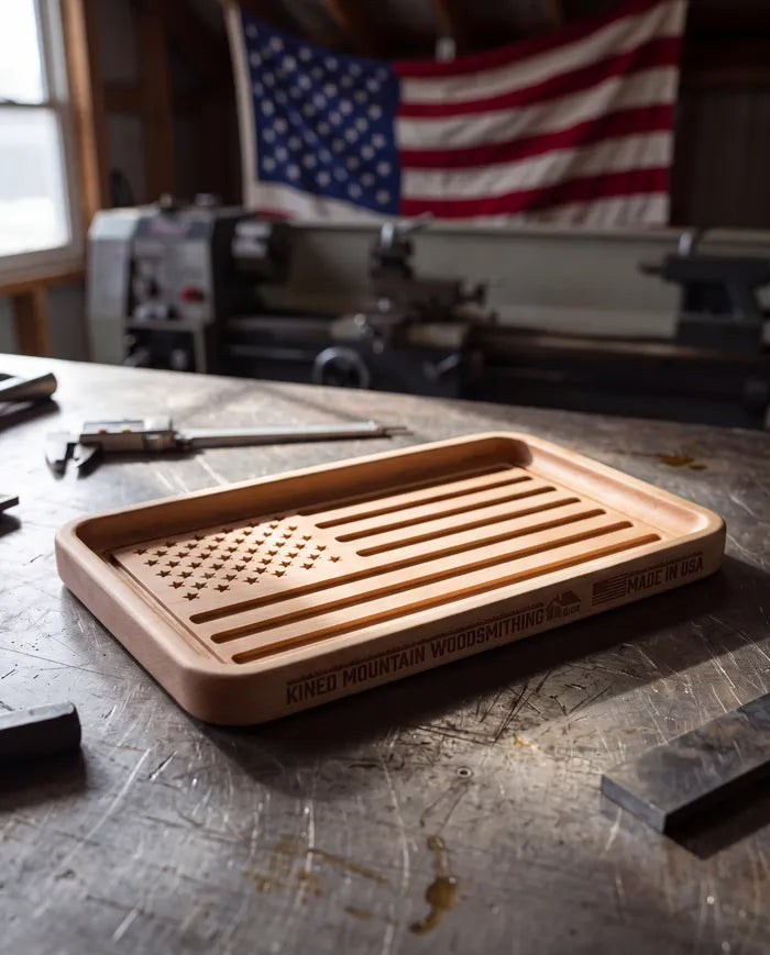 Hard maple American flag catchall tray on a steel workbench with a flag hanging in the shop behind.