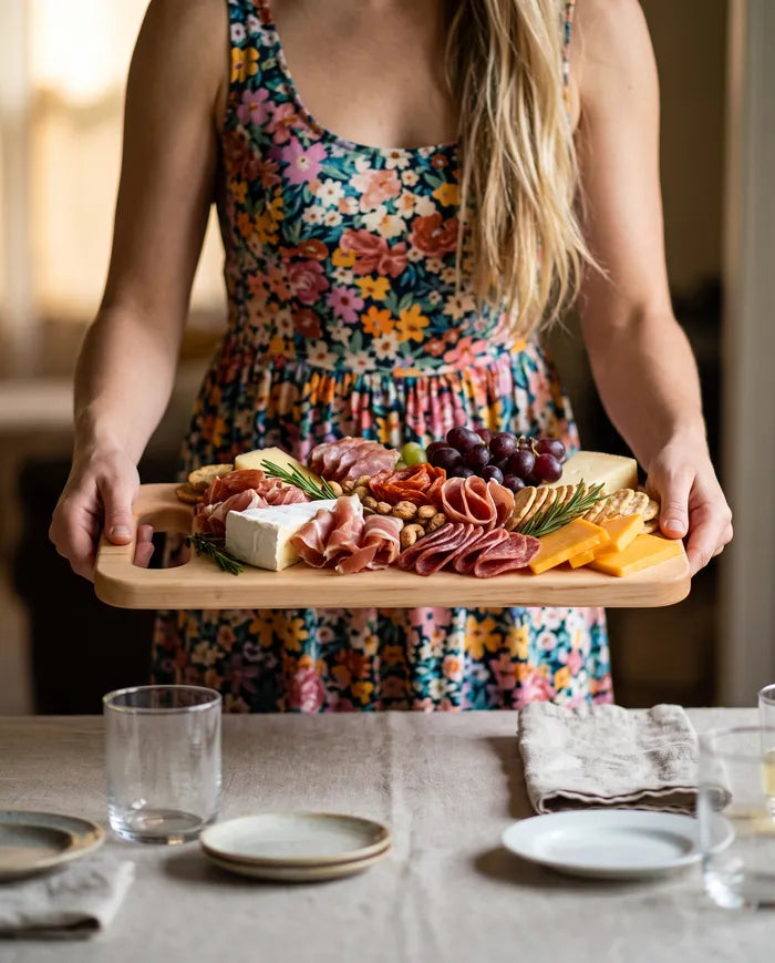 Woman carrying a maple bread board loaded with charcuterie, cheese, and grapes to a set table.