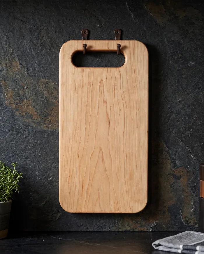 Hard maple bread board hanging by its handhold from two hooks on a dark slate kitchen wall.