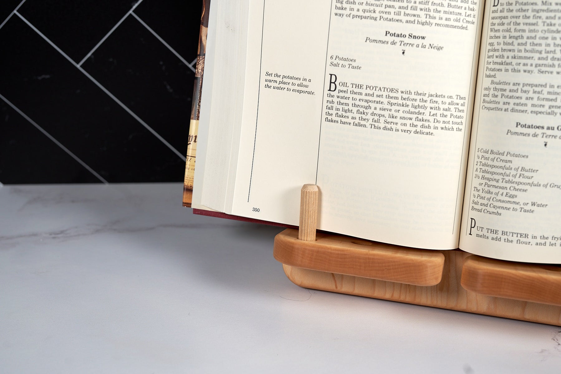 Maple cookbook stand holding an open cookbook on white marble kitchen counter with black tile backsplash.