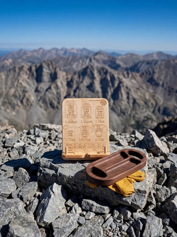 Maple cookbook stand with conversion chart and walnut serving tray displayed on rocks with scenic mountain view.