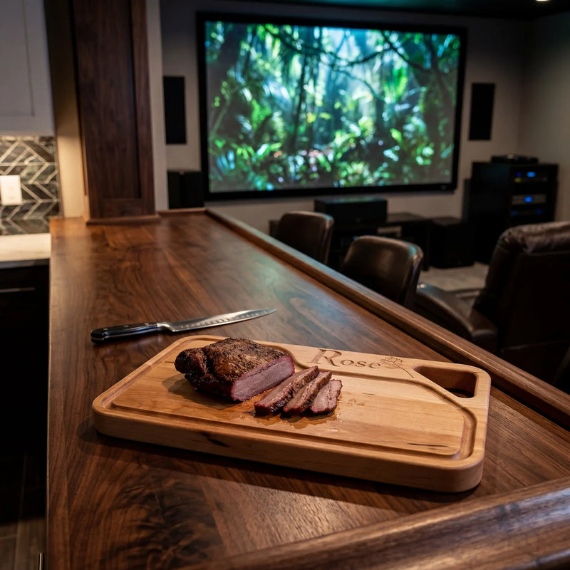 Maple engraved cutting board with sliced brisket and knife on bar counter in luxury home theater with large screen.