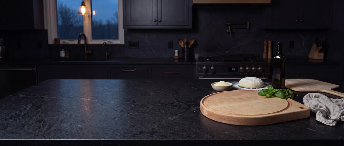 Maple round pizza cutting board on a dark stone kitchen island with pizza dough, basil, and olive oil ready to go.