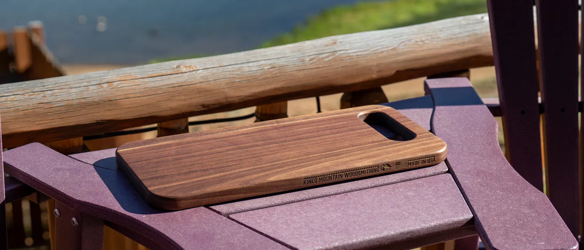 Walnut bread board with handhold cutout resting on the arm of an Adirondack chair on a lakeside deck.