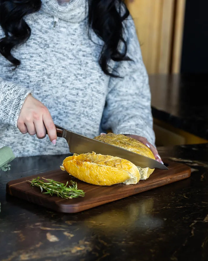 Slicing a rosemary loaf with a chef's knife on a walnut bread board on a black granite counter.