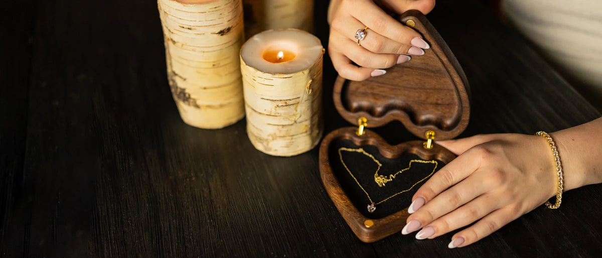 Hands opening a walnut heart-shaped keepsake box with a gold necklace inside, beside birch candles on a dark table.