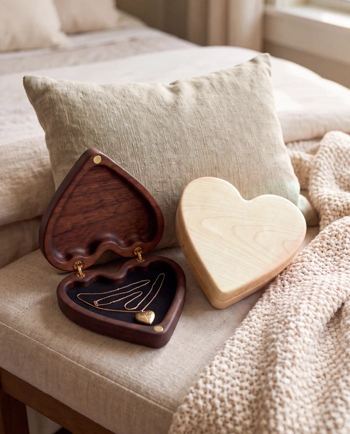 Open walnut heart keepsake box with a necklace on the felt beside a closed maple box, on a cushioned bench with a knit throw.
