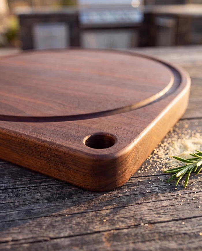 Close view of the juice groove and squared grip-hole corner on a walnut round pizza cutting board, on an outdoor table.