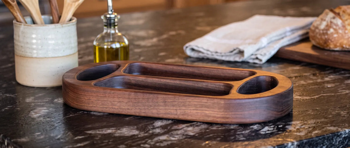 Empty walnut divided serving tray with two chambers and oval handholds on a dark granite kitchen counter beside a utensil crock and olive oil.