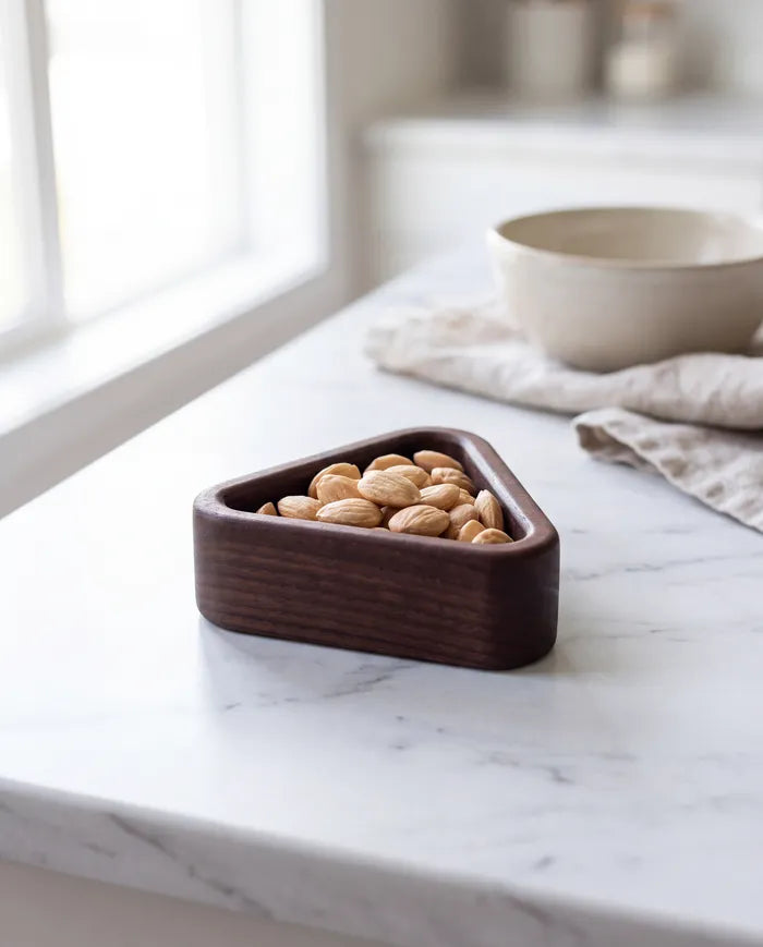 Triangular black walnut snack tray holding pistachios on a white marble kitchen counter.