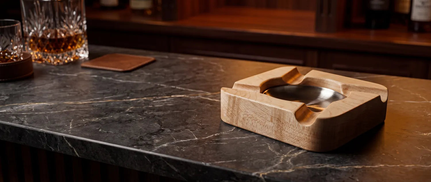 Handmade wood cigar rest on a bar counter with whiskey in a warm, low-lit setting.
